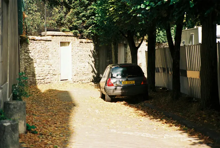 Ruelle bordée de murs en pierre, avec une porte fermée et une voiture garée sous les arbres